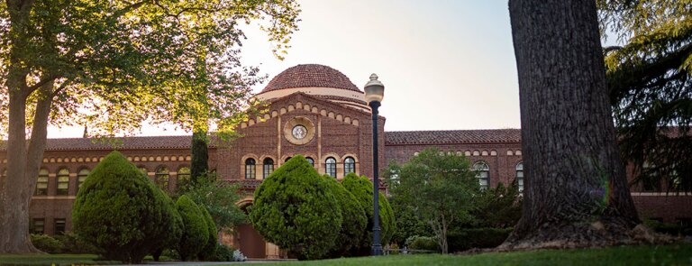 View of Chico State campus lawn with cone-shaped pine trees and brick building with rotunda in background